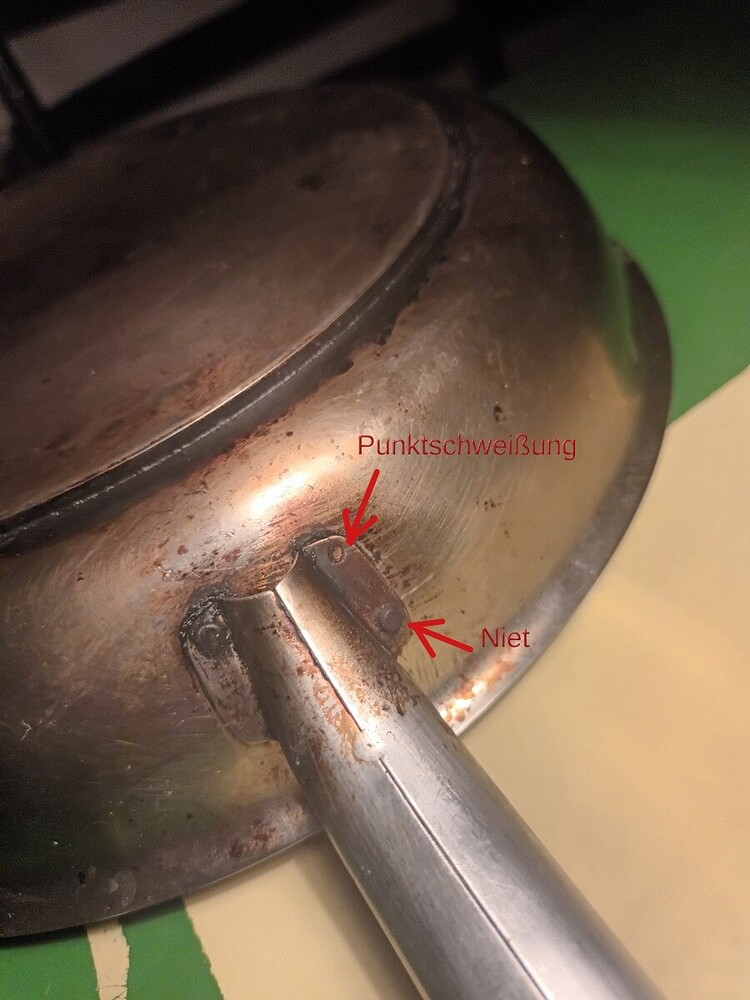 Nahaufnahme einer Bratpfanne. Zu sehen ist die Stelle, an der der Stil mit der Pfanne verbunden ist. Zwei rote Pfeile kennzeichnen die Stellen, wo eine Schweissung zu sehen ist und eine, wo eine Niete zu sehen ist.