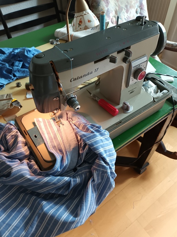 Eine Nähmanschine mit aufgelegtem Stoff und Kleidungsstücken darum herum.