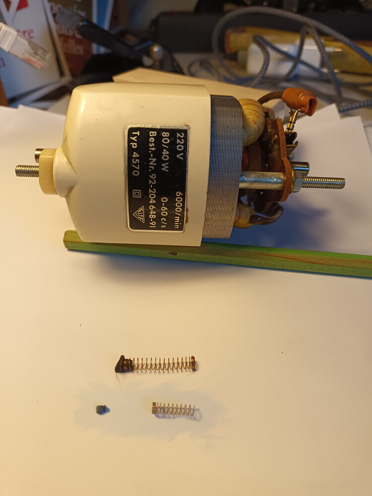 Nahaufnahme des ausgebauten Motors der Nähmaschine. Darunter liegen eine Spirale und ein sher kleines Stück Kohle.